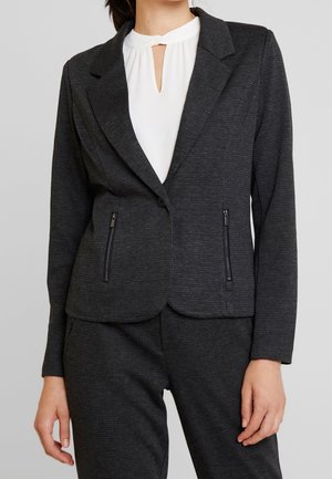 Woman wearing a dark gray checked blazer with zipper pockets over a white blouse with a keyhole neckline.