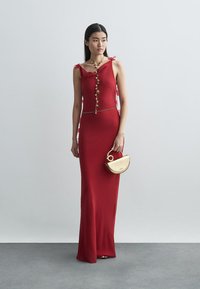 Κόκκινο εφαρμοστό φόρεμα με λεπτές τιράντες, διακοσμητικά κουμπιά και ασημένια ζώνη. Το μοντέλο κρατά μία μικρή χρυσή τσάντα.