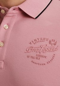 Polo rose avec un tissu texturé, comportant trois boutons, un col noir et blanc, et un logo en relief au centre.