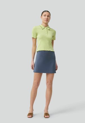 Kvinde står iført en lysegrøn kortærmet poloshirt med knapper, marineblåt mininederdel og brune åbne hælede sandaler mod grå baggrund.