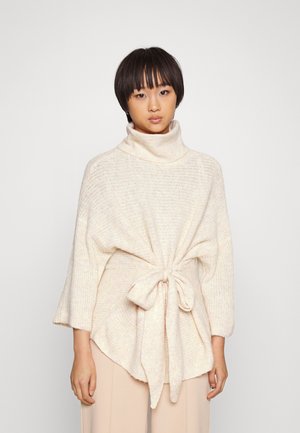 Cream-colored, oversized knit sweater with a turtleneck, open sleeves, and a tied waist detail. Soft texture and loose, flowing shape.