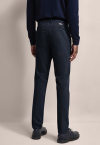 Donkerblauwe broek van een gestructureerde stof, met een platte tailleband, zijzakken en een strakke uitstraling, gecombineerd met zwarte schoenen.