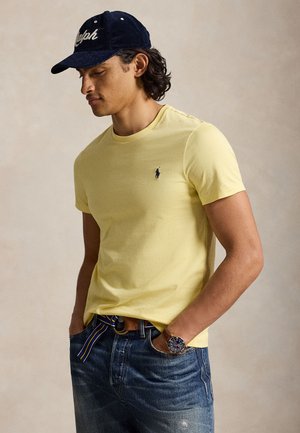 Homme aux cheveux bouclés foncés portant une casquette bleu marine, une chemise jaune clair à manches courtes avec un petit logo, un jean bleu, une ceinture rayée et une montre-bracelet, les mains dans les poches.