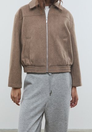 Femme portant une veste en laine marron zippée avec poches et taille élastique, associée à un pantalon de jogging gris à cordon de serrage.