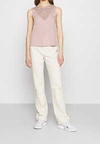 Różowy bezrękawnik z misternymi koronkowymi detalami przy dekolcie, w zestawieniu z białymi spodniami o prostym kroju i białymi trampkami. Całość o gładkiej fakturze.