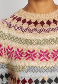 Beżowy sweter z dzianiny w wzorzyste paski w kolorach burgundu, różu, zieleni, bieli i czerni, z geometrycznymi wzorami oraz wzorami w kształcie śnieżynki.