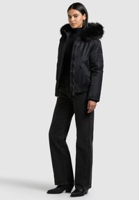 Geacă neagră puf cu glugă bordurată cu blană, prevăzută cu închidere cu fermoar, buzunare laterale și manșete rib, asortată cu blugi negri cu talie înaltă și cizme.