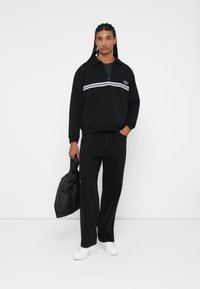 Sudadera negra con cuello con cremallera, raya horizontal blanca, pantalones negros a juego y zapatillas blancas. El modelo sostiene un bolso negro acolchado.
