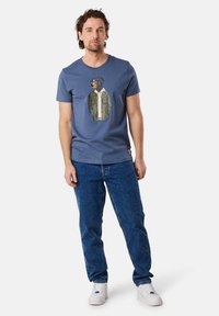 Short-sleeve blue t-shirt featuring a graphic of a dog in a green jacket, paired with blue jeans and white sneakers.