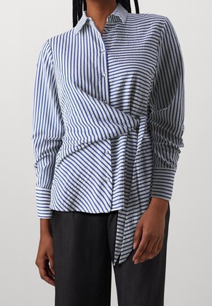 Personne portant une chemise boutonnée à rayures verticales bleues et blanches avec un nœud asymétrique noué à la taille, associée à un pantalon foncé.