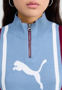 Blå strikkegenser med røde detaljer, full-zip krage, med en hvit hoppende katt-logo og vertikale striper langs skuldrene.