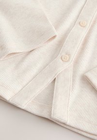 Chemise beige clair côtelée avec une fermeture boutonnée sur le devant, présentant une texture douce, un col arrondi et des manches longues.