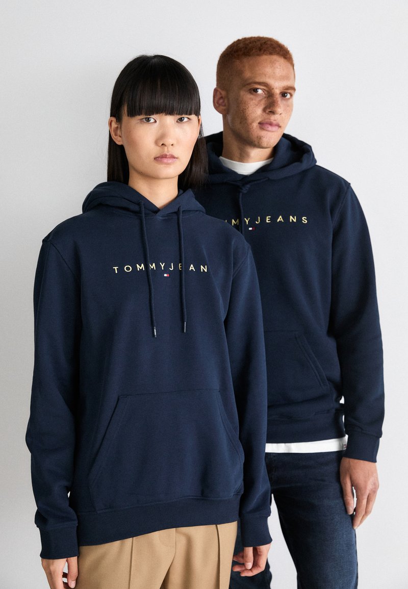 Des sweats à capuche bleu marine avec cordons et poches avant, arborant un logo doré "TOMMYJEANS". Le tissu semble doux avec une texture lisse.