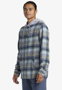 Das knöpfbare Flanellhemd hat ein blaues, grünes und gelbes Karomuster, eine graue Kapuze und eine Taschenklappe, gefertigt aus weichem Stoff.
