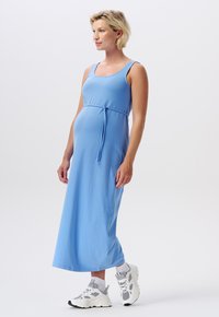 Robe maxi bleue avec un décolleté carré, design sans manches et un lien ajustable à la taille. Portée avec des baskets de sport blanches et grises.