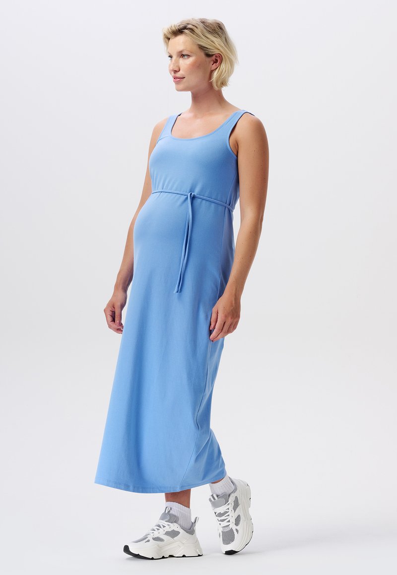 Robe maxi bleue avec un décolleté carré, design sans manches et un lien ajustable à la taille. Portée avec des baskets de sport blanches et grises.