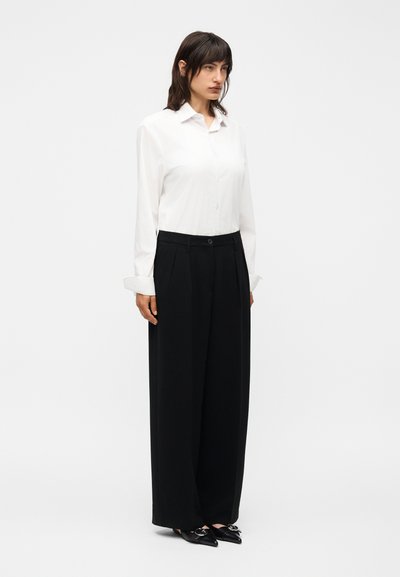 Γυναίκα στέκεται φορώντας ένα λευκό πουκάμισο με κουμπιά και μακριά μανίκια, μαύρο παντελόνι με φαρδιά πατζάκια και μαύρα μυτερά παπούτσια με ασημένιες αγκράφες.