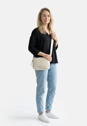 Kvinde iført sort top, lyseblå jeans, hvide sneakers, bærende en cremefarvet crossbody taske med justerbar rem, stående mod en hvid baggrund.