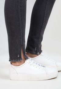 Vaqueros skinnies de denim negro con dobladillos deshilachados en los tobillos, combinados con zapatillas bajas blancas de textura suave y diseño simple.