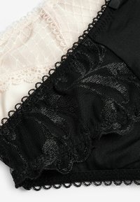 Lingerie nera e nuda in pizzo con bordi ondulati, caratterizzata da intricati motivi floreali su tessuto nero e una delicata texture a rete su nudo.