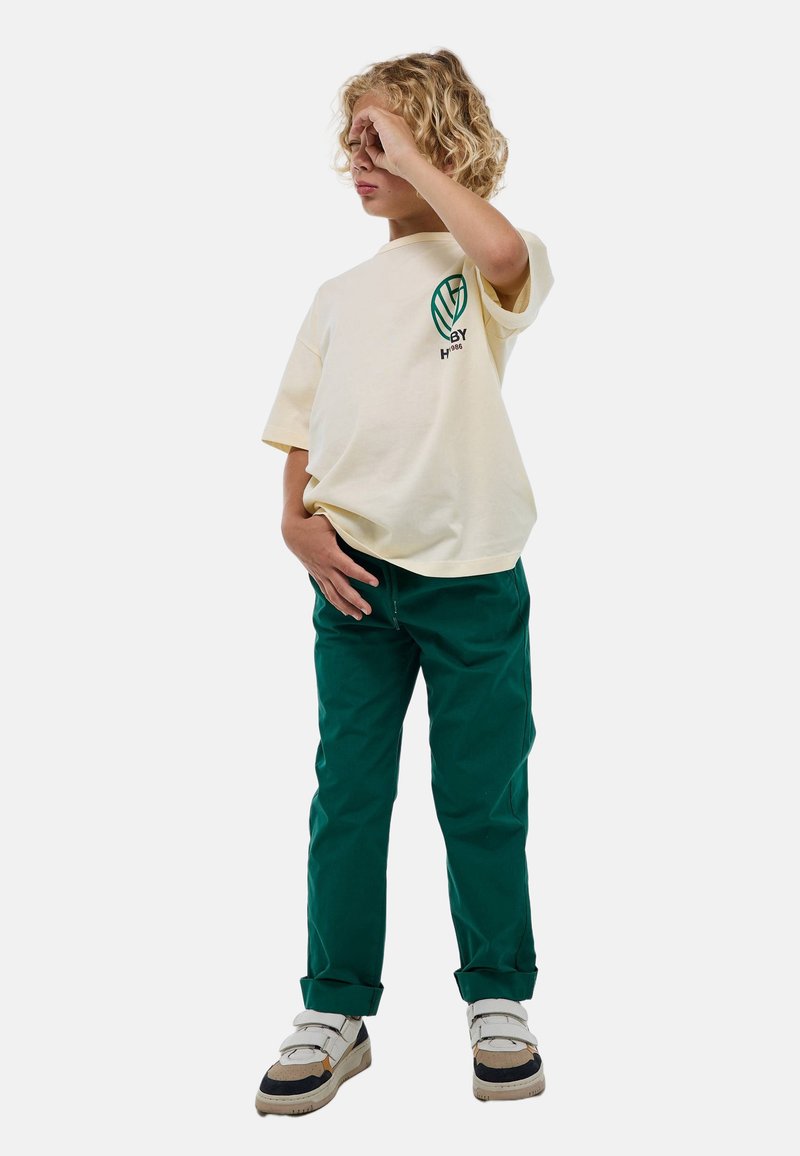 T-shirt à manches courtes crème avec logo vert, associé à un pantalon vert foncé et des baskets blanches avec des accents gris, noir et beige.