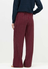 Osoba w granatowym swetrze oraz szerokich, bordowych spodniach z wysokim stanem i białymi pionowymi paskami, stoi z jedną ręką na biodrze.