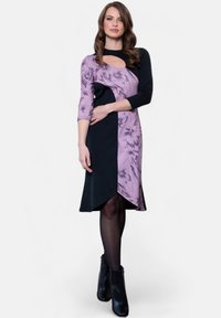 Violetti ja musta mekko kukkakuviolla, pitkät hihat, epäsymmetrinen helma ja avokolo kaula-aukossa. Mustat korkokenkäsaappaat.