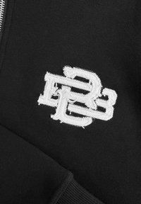 Zwarte stof met geborduurd wit logo met overlappende letters "D" en "B." Sterke textuur en geribbelde boorddetails.