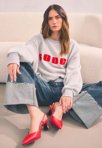 Grauer Sweatshirt mit "BOOCH" in rotem Text, kombiniert mit weiten Jeans aus Denim mit ausgefransten Säumen. Rote spitze Slingback-Schuhe mit Blockabsatz.