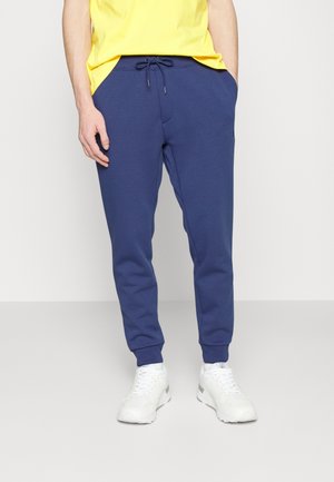 Homme portant un pantalon de jogging bleu avec cordon, un t-shirt jaune et des baskets blanches, debout devant un fond clair uni.