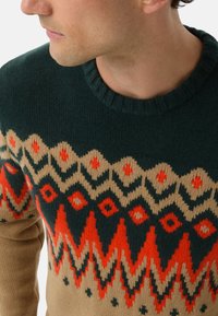 Man wearing a knitted sweater with zigzag and diamond patterns in dark green, beige, and bright orange colors, facing left.