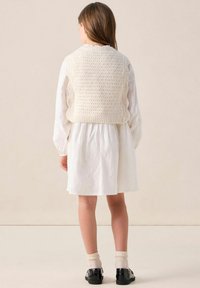 Strickweste in Creme mit strukturiertem Muster über einem weißen Langarmkleid; verfügt über einen gerafften Taillenbereich, knielangen Saum und Kniestrümpfe mit Schuhen.