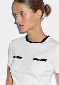 Witte kortekleurige top met zwarte afwerking en twee borstzakken met zilveren knopen. Soepele textuur met een getailleerd ontwerp.