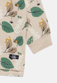 Tissu en coton beige clair avec des motifs de feuilles vertes, d'insectes et de fleurs. Présente un poignet en côtes et une étiquette "HUST et CLAIRE".