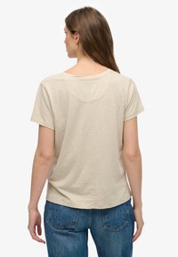 Beige Baumwoll-T-Shirt mit kurzen Ärmeln, legerer Passform, rundem Ausschnitt und dezenten Nahtdetails auf der Rückseite. Kombiniert mit blauen Jeans.