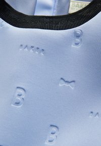 Svetle modrá tkanina s reliéfním textem "BAKER" a motivem mašle, s černým žebrovaným výstřihem. Hladká textura, moderní design.