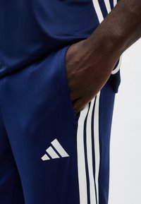 Άτομο που φοράει μπλε σκούρο παντελόνι Adidas με λευκές ρίγες και λογότυπο, το χέρι χαλαρά μέσα στην τσέπη.