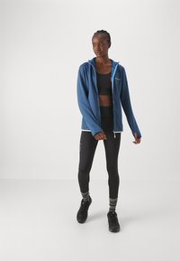 Modra zip-up kapuca iz mehkega materiala, v kombinaciji s črnimi legicami in temnimi čevlji. Obleka ima ozko prileganje in kontrastne akcentne dele.