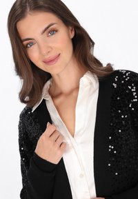 Schwarzer Cardigan mit Paillettenverzierungen auf einer Seite, kombiniert mit einem weißen Hemd mit Kragen. Weiche Textur, lockere Passform und lässiges Design.