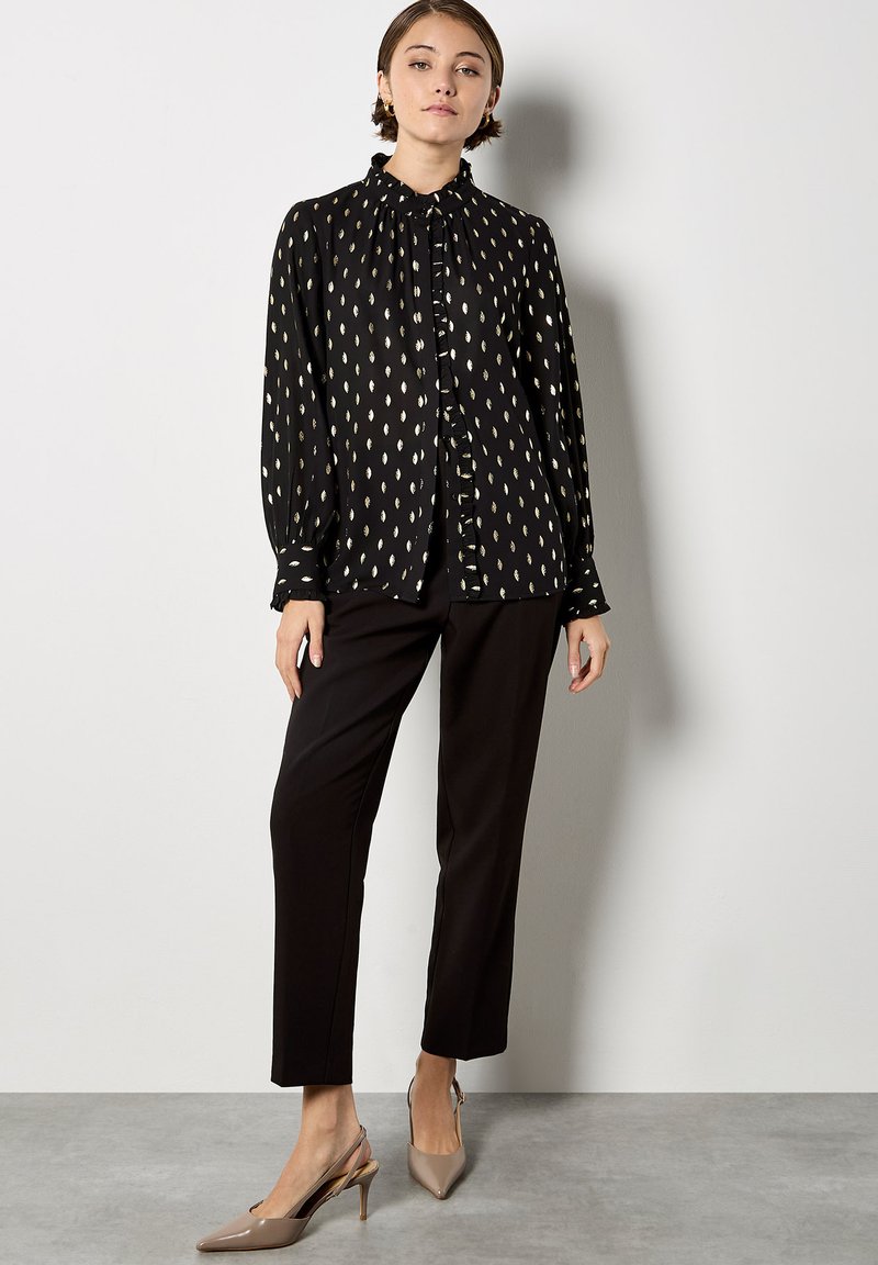 Black blouse with white leaf pattern, ruffled neckline, button-down front, and long sleeves. Paired with black trousers and nude slingback heels.