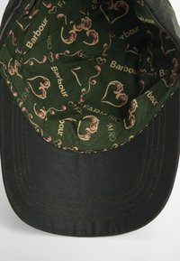 Black cap with a firm brim; interior features a green, patterned lining with flowers and logos in pink and gold tones.