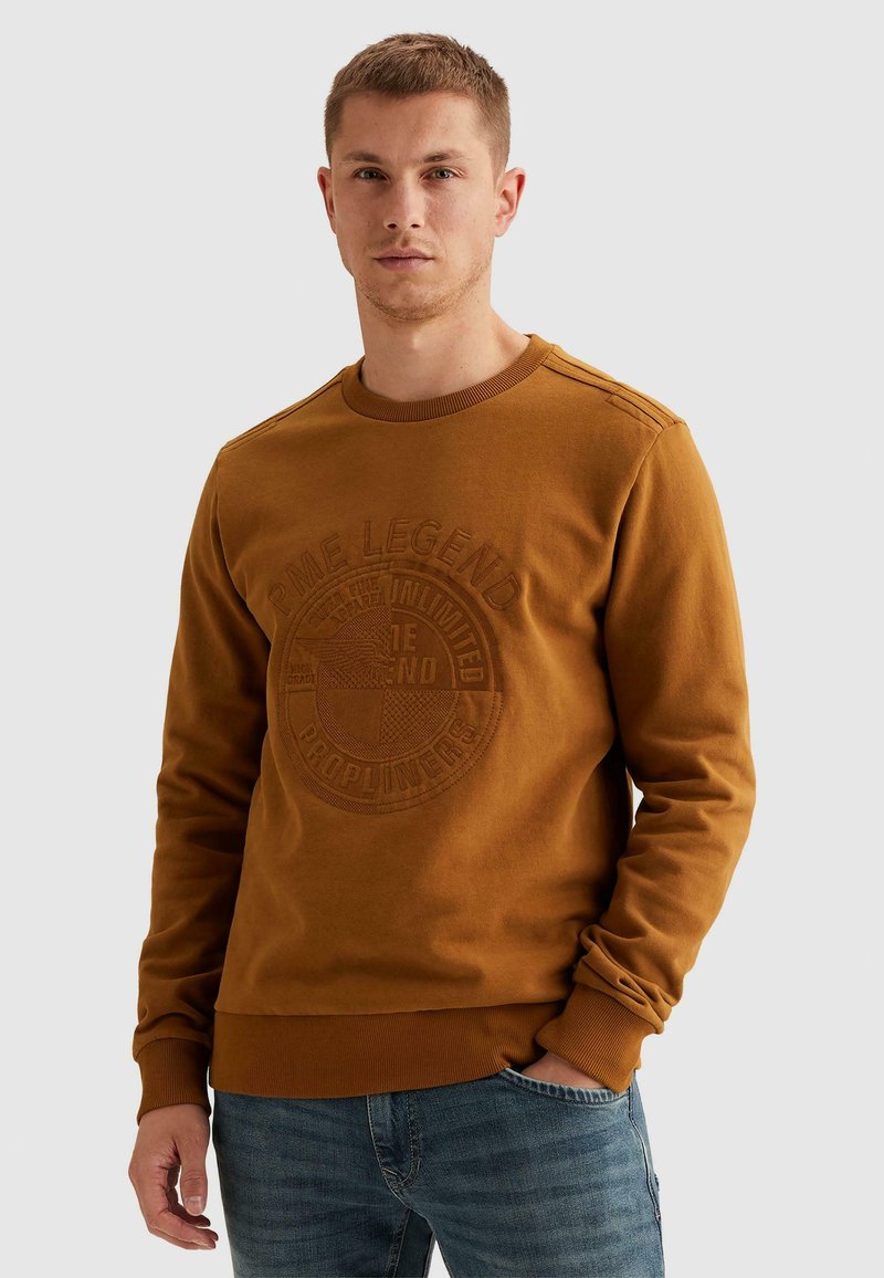 Jeune homme portant un sweat-shirt marron avec un logo circulaire en relief et du texte, associé à un jean bleu, debout devant un fond uni.