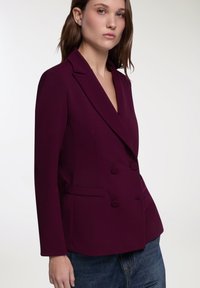 Blazer doppiopetto in un profondo viola, con rever a punta, dettagli con bottoni e tasche laterali, abbinato a jeans in denim.