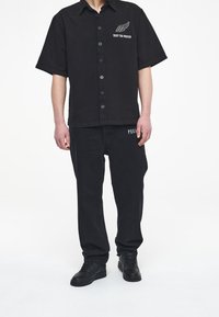 Svart kortärmad skjorta med knappar och framtryckt grafik, tillsammans med svarta raka jeans. Båda plaggen har en matt yta.