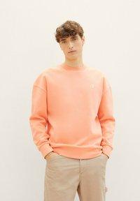 Koronkowy sweter z okrągłym dekoltem, ściągaczami i małym białym logo na piersi. Materiał wydaje się gładki i luźny.