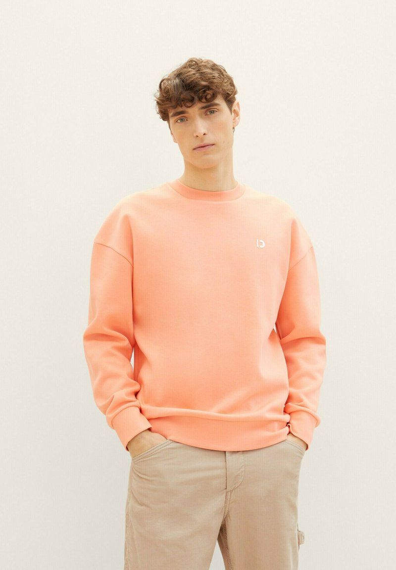 Koronkowy sweter z okrągłym dekoltem, ściągaczami i małym białym logo na piersi. Materiał wydaje się gładki i luźny.