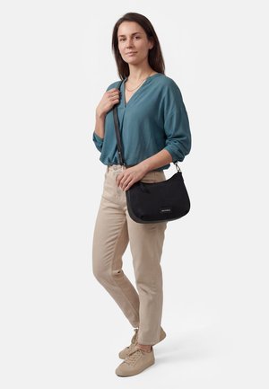 Frau trägt eine türkisfarbene Bluse, beige Hose und Turnschuhe, steht mit einer schwarzen Schultertasche vor einem schlichten weißen Hintergrund.