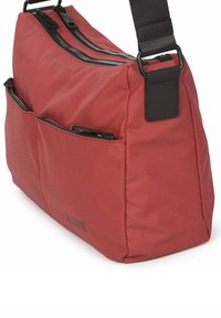 Bolso de hombro de tela roja con dos compartimentos con cremallera, bolsillos exteriores y una correa negra ajustable. Presenta un detalle de logotipo en el lateral.