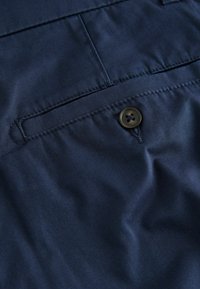 Tissu bleu foncé avec une poche arrière cousue et un bouton noir à quatre trous qui maintient le rabat de la poche.