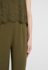 Top en dentelle verte à motif floral associé à un pantalon vert uni assorti, montrant le bas du torse et le bras droit d'une personne.
